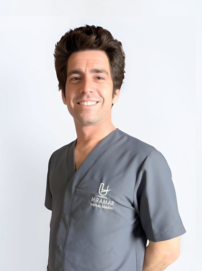 Dr.A Cuevas Dr. Alberto Cuevas, responsable del Área de Cirugía Maxilofacial de Instituto Médico Miramar, de cuerpo entero sonriente con uniforme sanitario gris